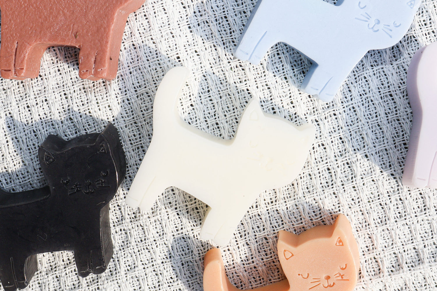 A set of cat-shaped soaps in various pastel and neutral colors, including black, white, light blue, lavender, peach, and terracotta, arranged on a textured white linen fabric.