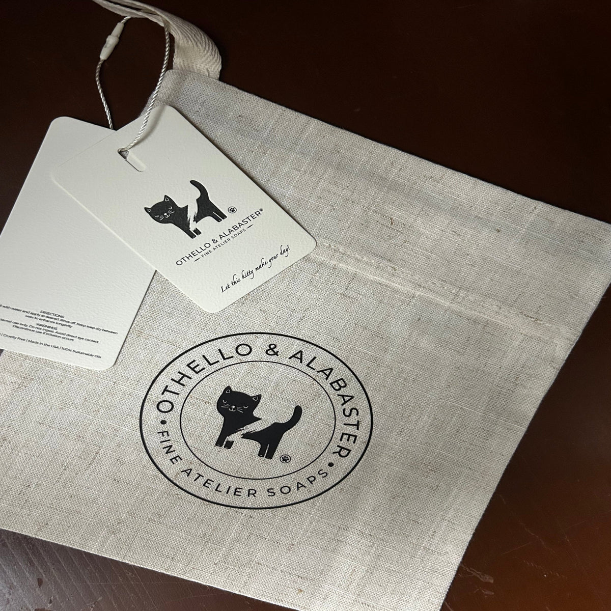 A light beige linen pouch with a black circular logo featuring a cat and the text 'Othello & Alabaster Fine Atelier Soaps', shown with a white paper hangtag on a dark brown wooden surface.