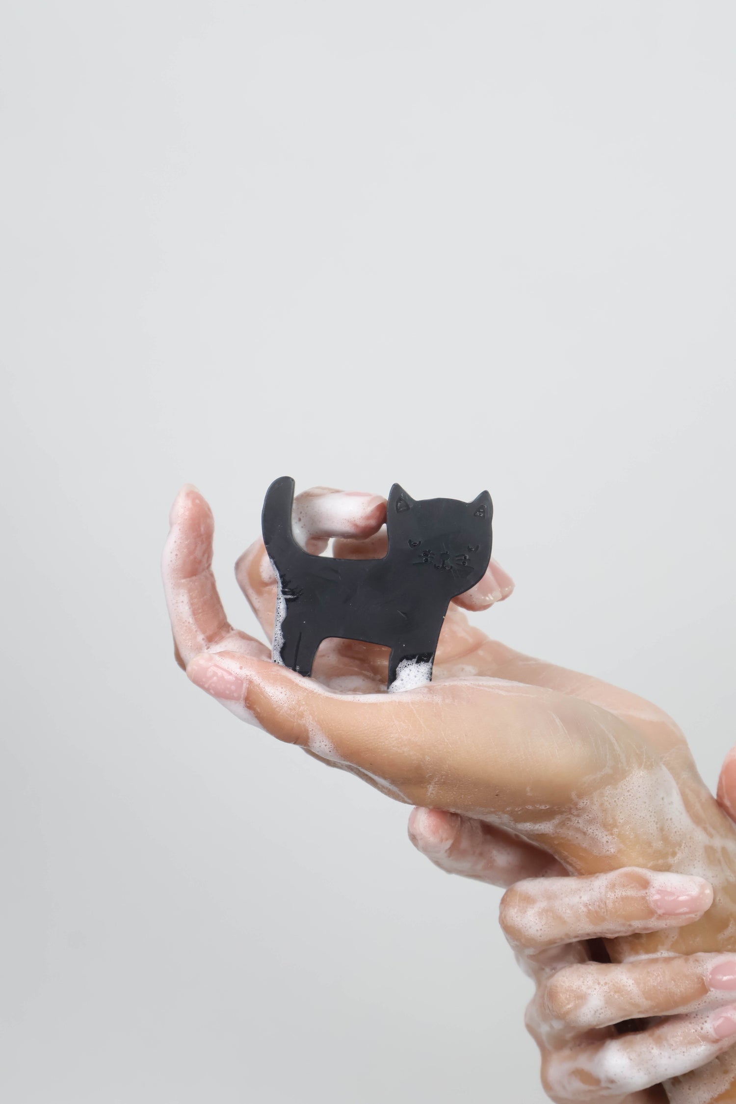 A small black cat-shaped soap held between two hands covered in white soap foam against a plain light gray background.